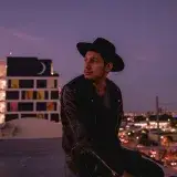 Stylish man in black leather jacket and hat sits on Miami rooftop at evening twilight with city lights.