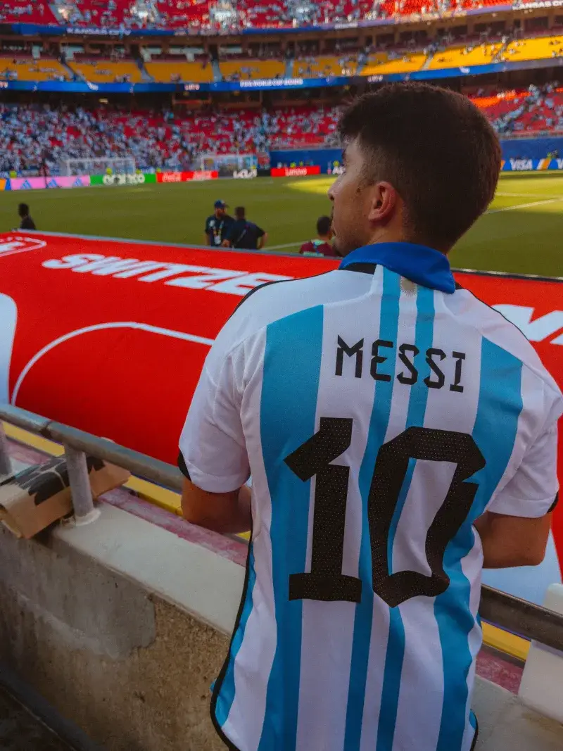 Fan in Argentina jersey with 'Messi 10' watching football match at crowded stadium.