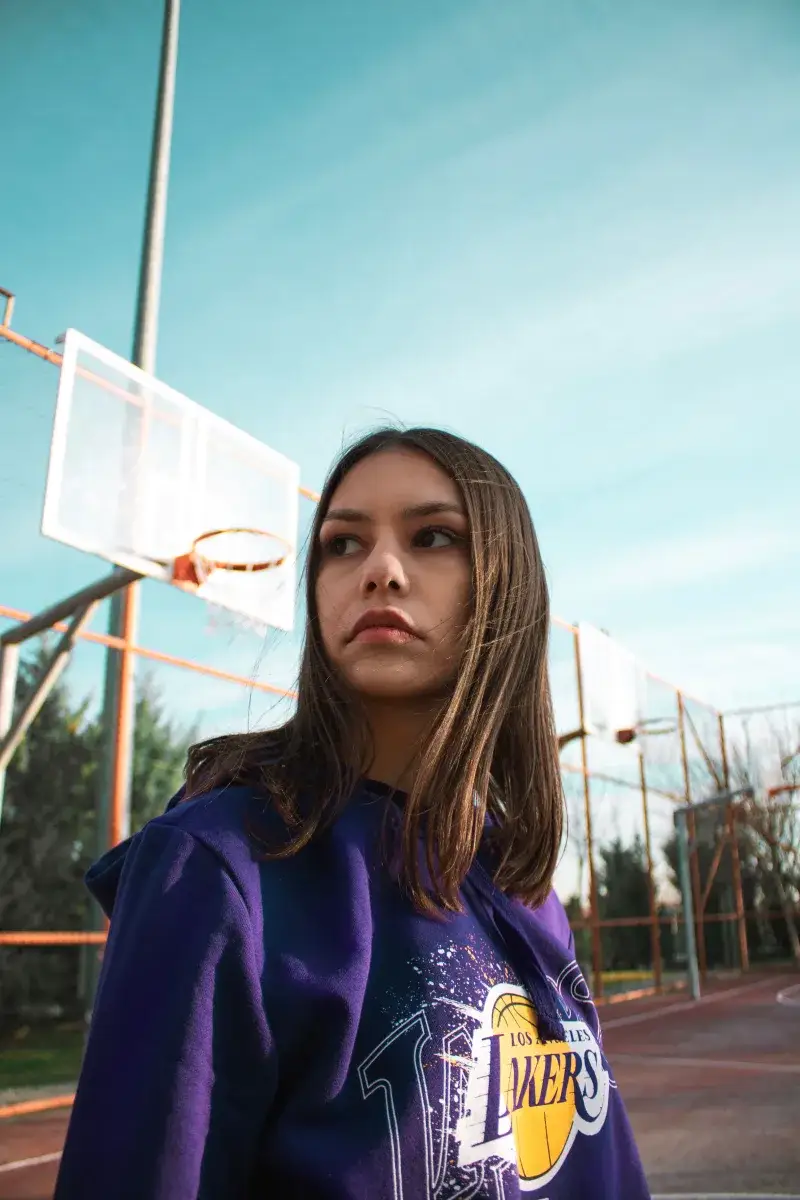 Portrait of a woman in a Lakers hoodie on an outdoor basketball court under a clear sky.