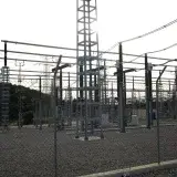 Outdoor electrical power substation with high voltage equipment and safety signs, surrounded by fencing.