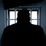 Dark silhouette of a man in a prison cell, backlit by window bars.