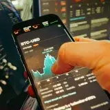 Una mano sostiene un smartphone que muestra un gráfico de BTC-USD entre pantallas de trading digital, destacando las finanzas de criptomonedas.