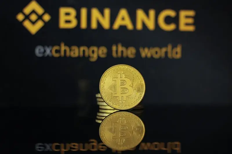 Primer plano de una moneda de Bitcoin con el logo de Binance y texto reflejado en una superficie oscura.