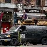 Man on top of loaded van in a bustling New York City streetscape with discount store signage.