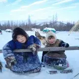 Two children enjoying a snowy winter day in the Yamal-Nenets region of Russia, surrounded by sleds and a dog.