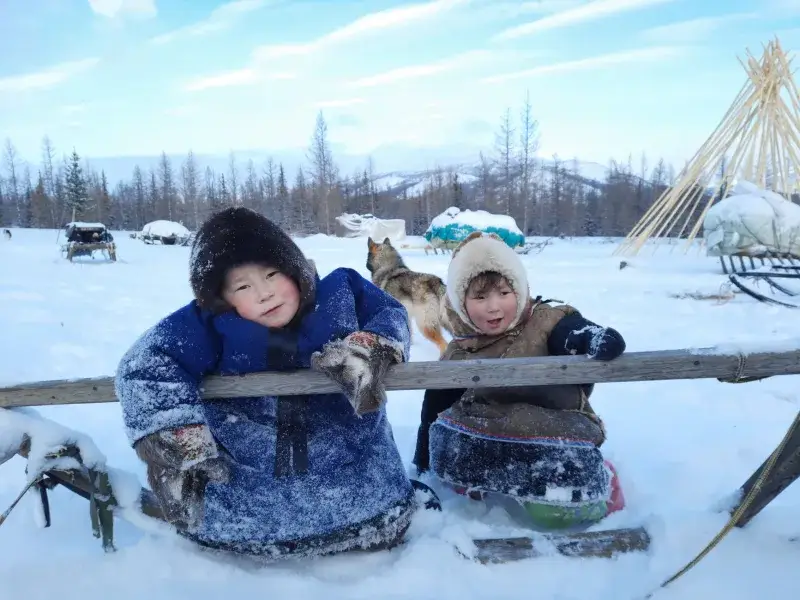 Two children enjoying a snowy winter day in the Yamal-Nenets region of Russia, surrounded by sleds and a dog.