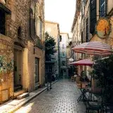Explore the picturesque, sunlit alleyway of Vence in Provence, lined with quaint cafes and stone buildings.