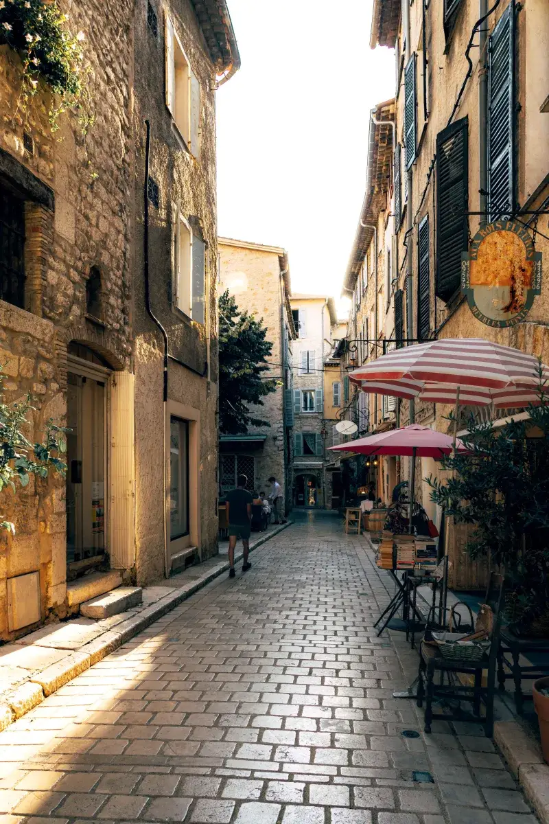 Explore the picturesque, sunlit alleyway of Vence in Provence, lined with quaint cafes and stone buildings.
