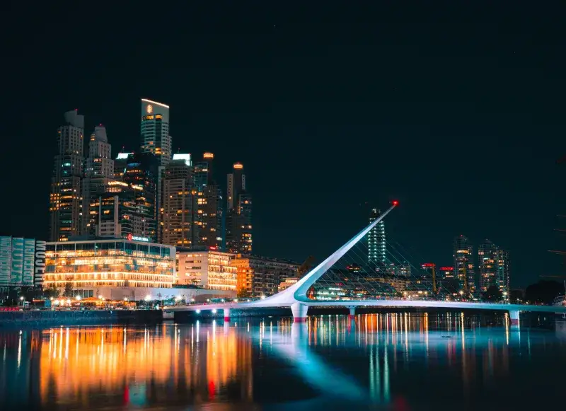 Modern architectural view of Puente de la Mujer and Buenos Aires skyline reflected in water at night.