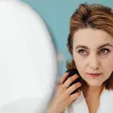 A woman styles her hair while looking into a mirror indoors, focusing on personal grooming.