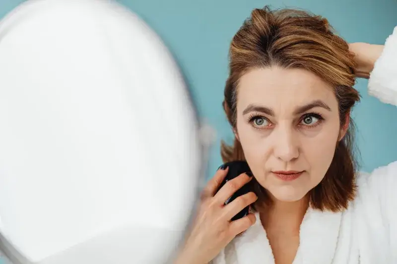 A woman styles her hair while looking into a mirror indoors, focusing on personal grooming.