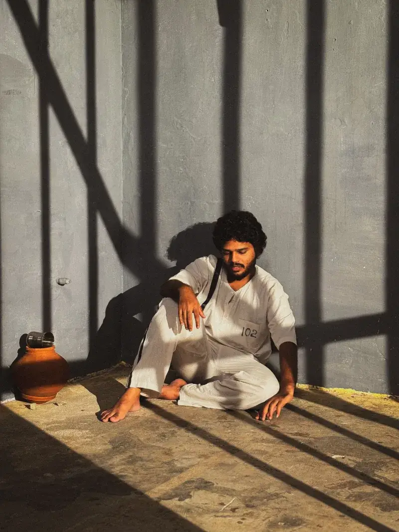 A man in a white uniform sits on a sunlit prison floor, casting shadows.