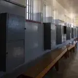 Empty prison cell block with benches and cabinets, lit by barred windows casting shadows.