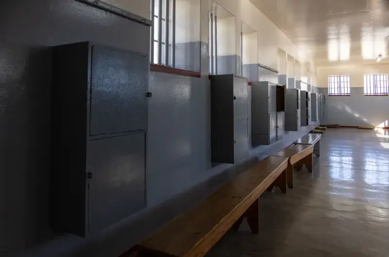 Empty prison cell block with benches and cabinets, lit by barred windows casting shadows.