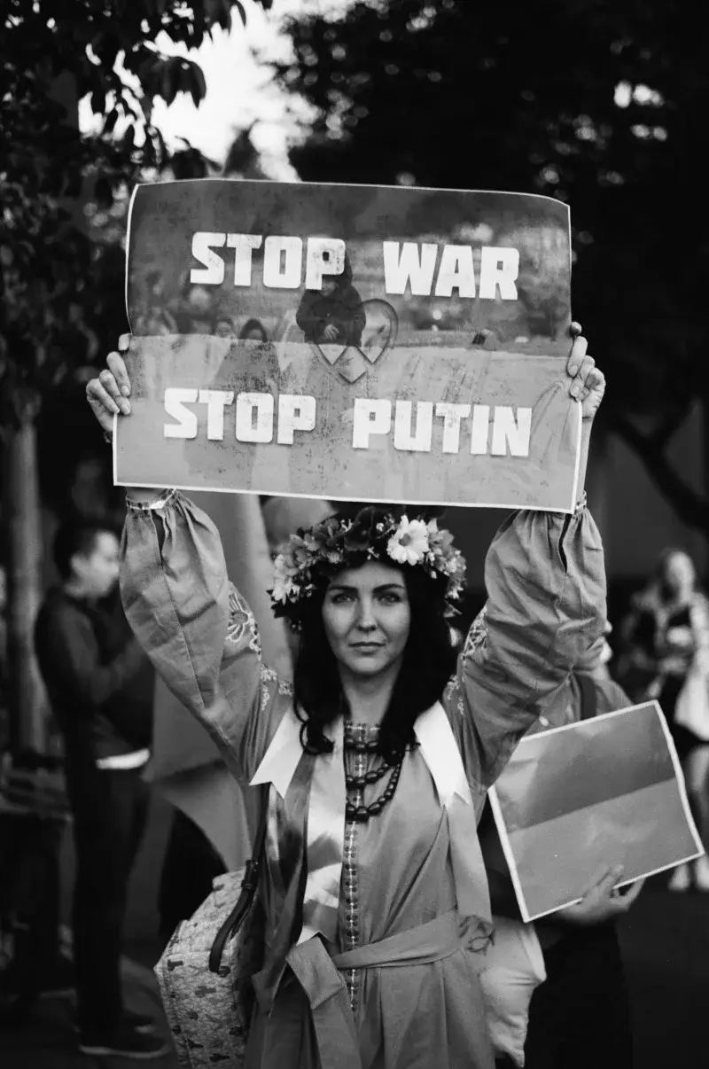 Black and white photo of a woman holding a banner reading 'Stop War, Stop Putin' during a protest.