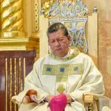 A clergyman dressed in ceremonial robes sits in a beautifully adorned church interior in Baliwag, Philippines.