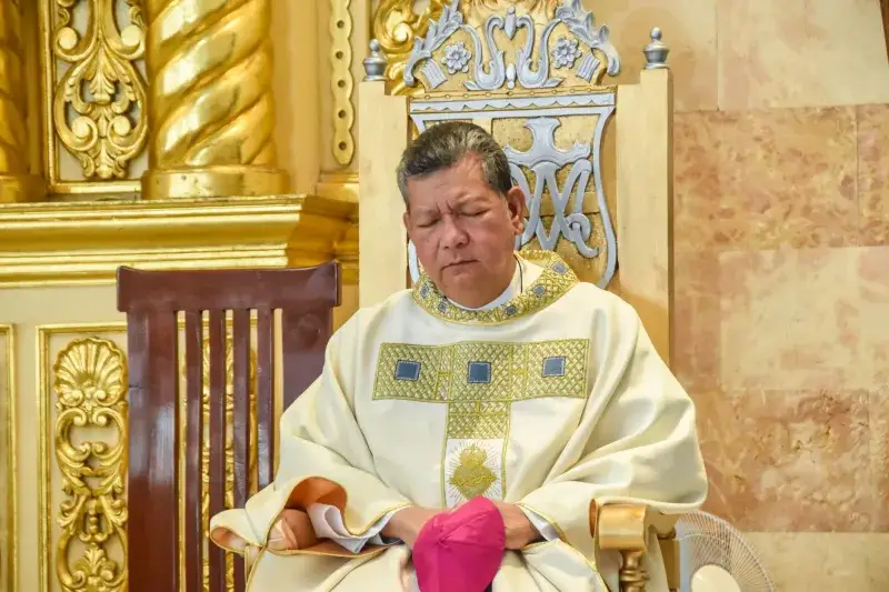 A clergyman dressed in ceremonial robes sits in a beautifully adorned church interior in Baliwag, Philippines.