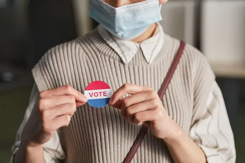 A person wearing a mask holds a voting badge, symbolizing participation in the election.
