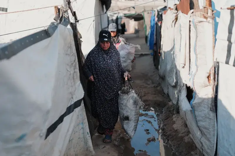 Two women navigate a narrow alley in a refugee camp, carrying bags. Real-life struggle captured.