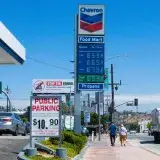 People walking near a Chevron gas station with prominent signage and parked vehicles on a sunny day.