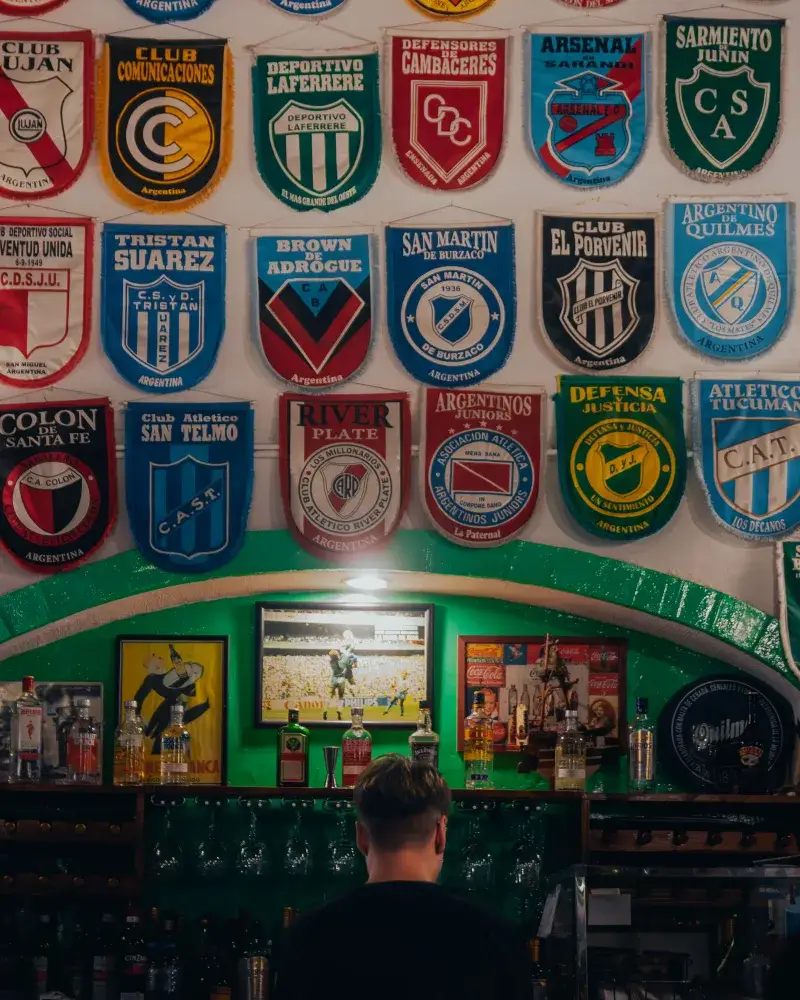 Una escena animada de un bar con logotipos coloridos de clubes de fútbol exhibidos en la pared.